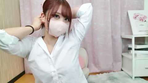 Media: Video of an East Asian woman with fair skin, light pink hair, and a white face mask, wearing a white button-up shirt, sitting at a desk with a pink background and white storage shelves.