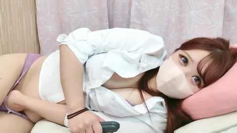 Media: Video of a young Asian woman with long brown hair, wearing a white button-up shirt, purple panties, and a face mask, lying on a pink pillow, in a soft-lit room.