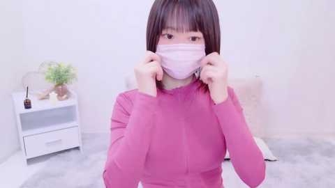 Media: Video of an Asian woman with straight, shoulder-length black hair, wearing a pink mask, pink long-sleeve shirt, and a white headband. She is adjusting her mask in a bright, minimalistic room with white walls and a gray carpet.