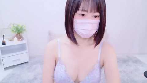 Media: Video of an Asian woman with a pale complexion and straight black hair, wearing a white lace bra and a pink surgical mask, sitting on a white carpet in a minimalist room.