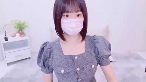 Media: Video of a young East Asian woman with straight black hair, wearing a gray button-up dress, white face mask, and standing in a bright, minimalistic room with white furniture and a potted plant.