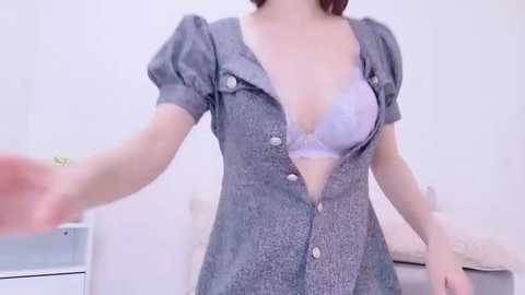 Media: Video of a fair-skinned woman in a low-cut, button-up, grey dress revealing a lavender bra, with blurred background of a white room and bed.