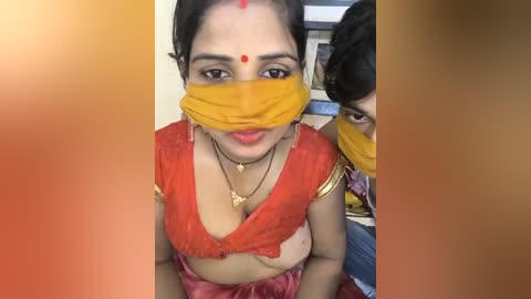 Media: Video of a woman with medium skin tone and dark hair, wearing a bright orange saree, red bindi, and yellow face mask, partially covering her face.