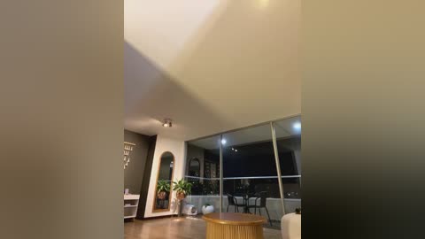 Media: Video of a modern, minimalist living room with a high ceiling, wooden floor, and large glass windows revealing an urban night skyline. A round wooden table and a potted plant are visible, with a black wall and a white arched doorway on the left.