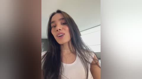 Media: A video of a young Asian woman with long black hair, wearing a white t-shirt, standing in a modern, minimalist room with white walls and a glass door.