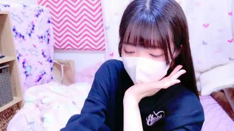 Media: Video of an Asian girl with straight black hair, wearing a black school uniform, a white mask, and a white headband, sitting on a bed with pink and white bedding, in a room with a red and white chevron-patterned wall.