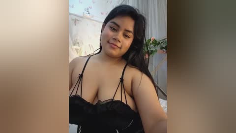 Media: A video of a plus-size woman with medium skin tone and long black hair, wearing a black lace lingerie top, standing indoors with floral wallpaper and a plant in the background.