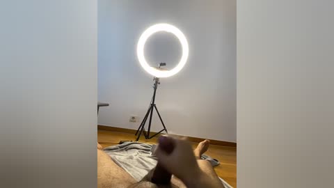 Media: Video of a man lying on a wooden floor, with a bright ring light and tripod in the background.