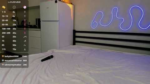 Media: Video of a modern bedroom with a white bed, a black headboard, a neon \"LUV\" sign on the wall, and a remote control on the bed, with an overlay of a baby monitor app showing a baby sleeping in a crib.