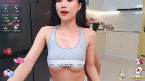 Media: Video of an Asian woman with long black hair, wearing a gray Calvin Klein sports bra, standing in a modern kitchen with white cabinets, a black fridge, and a digital clock displaying 15:15.