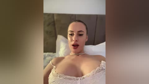 Media: Video of a fair-skinned woman with brown hair tied back, wearing a white lace bra, lying on a bed with a gray upholstered headboard. She has a surprised or shocked expression.