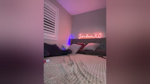 Media: A video of a dimly lit bedroom featuring a grey headboard, beige quilt, and pink neon sign reading \"Jordanna X0.\" The room has muted tones and a cozy, intimate atmosphere.