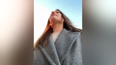 Media: Video of a woman with long, straight brown hair, wearing a gray, textured coat, standing outdoors with a clear blue sky background, looking upwards with a serene expression.