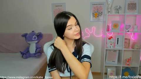 Media: Video of a young, fair-skinned woman with long black hair, wearing a sailor schoolgirl outfit, adjusting her hair in a cozy, pink-lit room with stuffed toys, framed pictures, and a neon sign.