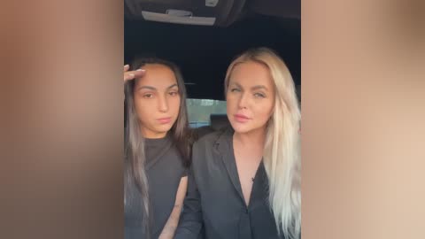 Media: Video of two women, one with long dark hair, one with long blonde hair, both wearing black blazers, inside a car, looking serious.