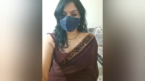 Media: Video of an Indian woman with medium skin tone, wearing a black mask, maroon saree with gold embroidery, and a red bindi. Background features a white wall and patterned fabric.