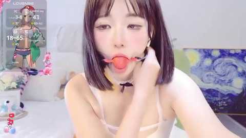 Media: A video of an Asian woman with straight black hair and fair skin, wearing a white bra, gagged with a red ball gag, in a bedroom with a Van Gogh painting and a pink flower arrangement in the background.