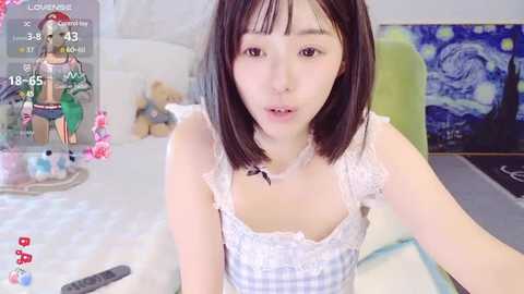 Media: Video of a young East Asian woman with straight black hair, wearing a white checkered top, sitting on a bed with a blue blanket. The background features a green chair and a colorful painting.