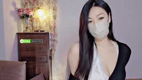 Media: Video of an Asian woman with long black hair, wearing a white lace bra and black top, standing in a room with a vintage wooden dresser and a lamp.