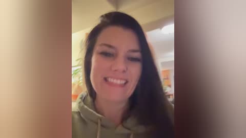 Media: Video of a smiling young woman with long dark hair, wearing a green hoodie, in a cozy indoor setting with blurred background details.