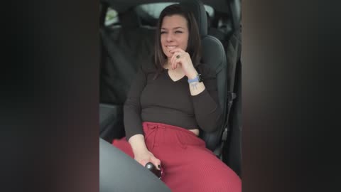 Media: Video of a woman with straight brown hair, wearing a black long-sleeve top and red pants, seated in a car, holding a phone, smiling, with a blurred background.