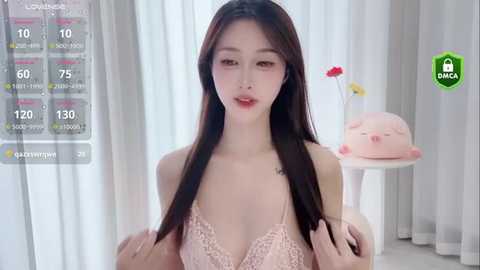 Media: Video of a young Asian woman with long black hair, wearing a pink lace bra, standing in a bright room with a cat statue and white curtains in the background.