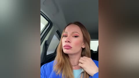 Media: A video of a Caucasian woman with light skin, straight blonde hair, wearing a blue jacket, sitting in a car with a neutral background. She has a neutral expression and is slightly puckering her lips.