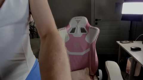 Media: Video of a woman in a white tank top, standing near a pink gaming chair with a white and blue logo, in a dimly lit room with a desk and monitor.