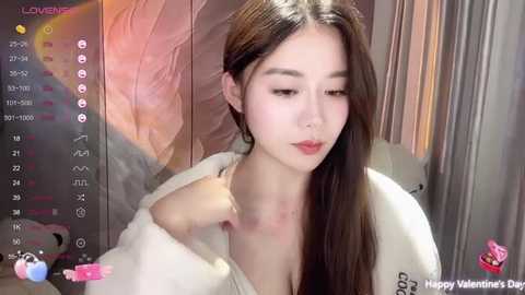 Media: Video of an Asian woman with long brown hair in a white robe, looking pensive, set against a blurred, pink-toned background. Text overlay displays \"LOVE\" and \"Happy Valentine's Day\" in pink.