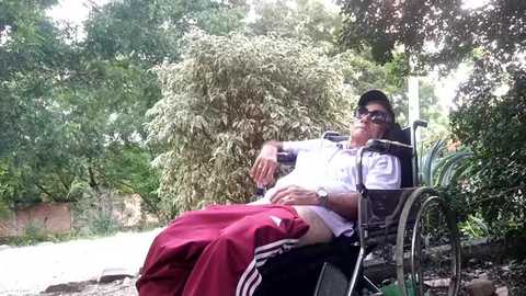 Media: Video of a man in a wheelchair with sunglasses, wearing a white shirt and red shorts, reclining outdoors under a tree, surrounded by lush greenery and a garden path.