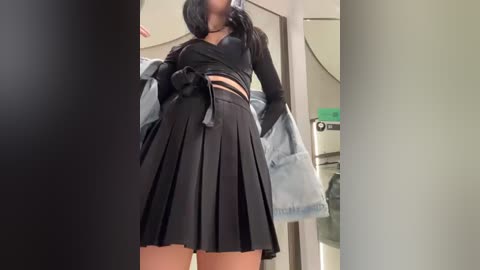 Media: Video of a young woman with long black hair, wearing a black crop top and high-waisted pleated black skirt, standing in a bathroom with a green sink and towel rack in the background.