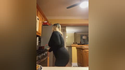 Media: Video of a blonde woman in a black dress cooking in a warm, cozy kitchen with wooden cabinets, stove, and a ceiling fan.