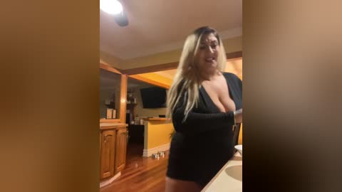 Media: A video of a curvy, light-skinned woman with long, blonde hair, wearing a low-cut black dress, standing in a cozy, warmly-lit kitchen with wooden cabinets and a flat-screen TV.