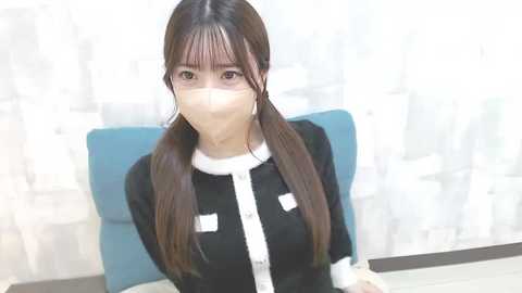 Media: Video of an Asian woman with long black hair in pigtails, wearing a black dress with white accents, sitting indoors with a blue chair and a blurred background.