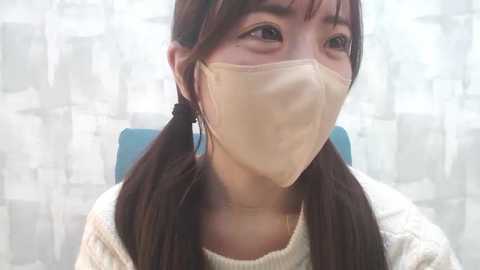 Media: Video of an Asian woman with long brown hair in pigtails, wearing a beige surgical mask and white sweater, seated in front of a white and gray abstract-patterned background.