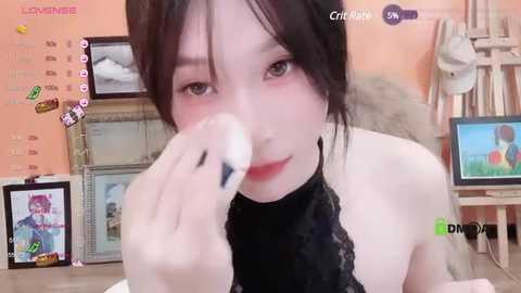 Media: A video of a young East Asian woman with fair skin, black hair, and light makeup, wearing a black lace top, holding a makeup brush, in a cozy, cluttered room with a TV showing a pink-haired anime character.