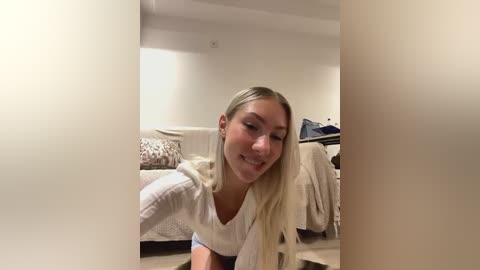 Media: Video of a smiling, fair-skinned, blonde woman in a white sweater, crouching in a dimly-lit, cozy living room with beige couch, blankets, and a blue bag.