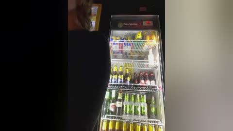 Media: Video of a well-stocked fridge with green, yellow, and brown bottles of various beverages, a fruit tray, and a person's hand reaching inside. The background is dark.