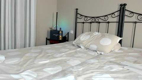 Media: Video of a neatly made bed with a cream-colored duvet featuring large white polka dots, a matching pillow, and a black wrought-iron headboard. A small black nightstand with a blue box, a lamp, and a glass on the left side.