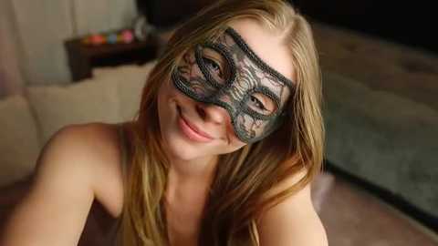 Media: A video of a smiling young woman with long blonde hair wearing a black lace masquerade mask, sitting indoors on a beige sofa, with a blurred background featuring a wooden table and a dark carpet.