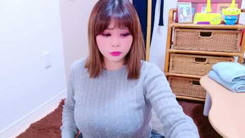 Media: Video of an East Asian woman with shoulder-length brown hair, pink lipstick, and a gray sweater, sitting on a brown carpet in a room with a wicker chest, colorful toys, and a white wall.