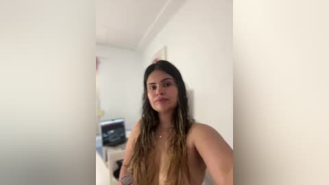 Media: Video of a young woman with long, wavy, brown hair, standing naked against a white wall in a narrow hallway, with blurred, indistinct objects in the background.