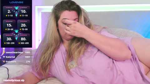 Media: Video of a plus-sized woman with long blonde hair, wearing a light purple robe, covering her face with her hand. Background shows a modern bedroom with white walls and a bed.