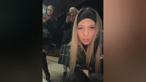 Media: Video of a young woman with long blonde hair and a black headband, wearing a black leather jacket, posing in a dimly lit bar with three other women in dark clothing in the background.