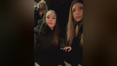 Media: Video of two young women, one with long brown hair and another with long blonde hair, wearing black coats, seated in a dimly lit room with another woman in the background.