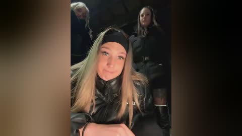 Media: Video of a young woman with long blonde hair, wearing a black leather jacket, seated in a dimly lit room. Background shows two other women in similar attire, one with short dark hair and another with long dark hair.