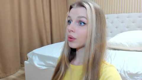 Media: Video of a young, fair-skinned woman with long blonde hair, wearing a yellow shirt, gazing thoughtfully at a neatly made bed with white sheets and a beige headboard in a softly lit room.