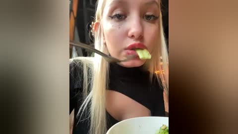 Media: Video of a young blonde woman with fair skin, wearing a black top, eating a leafy green salad with a fork, her lips slightly open, in a dimly lit indoor setting.