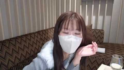 Media: Video of an Asian woman with long dark hair, wearing a white mask and light blue robe, lying on a brown and beige zigzag-patterned couch, indoors with vertical striped wallpaper and a lit candle on a side table.