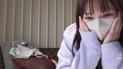 Media: Video of an East Asian woman in a white robe, holding her face, wearing a white mask, surrounded by a messy room with a patterned couch and vertical blinds.